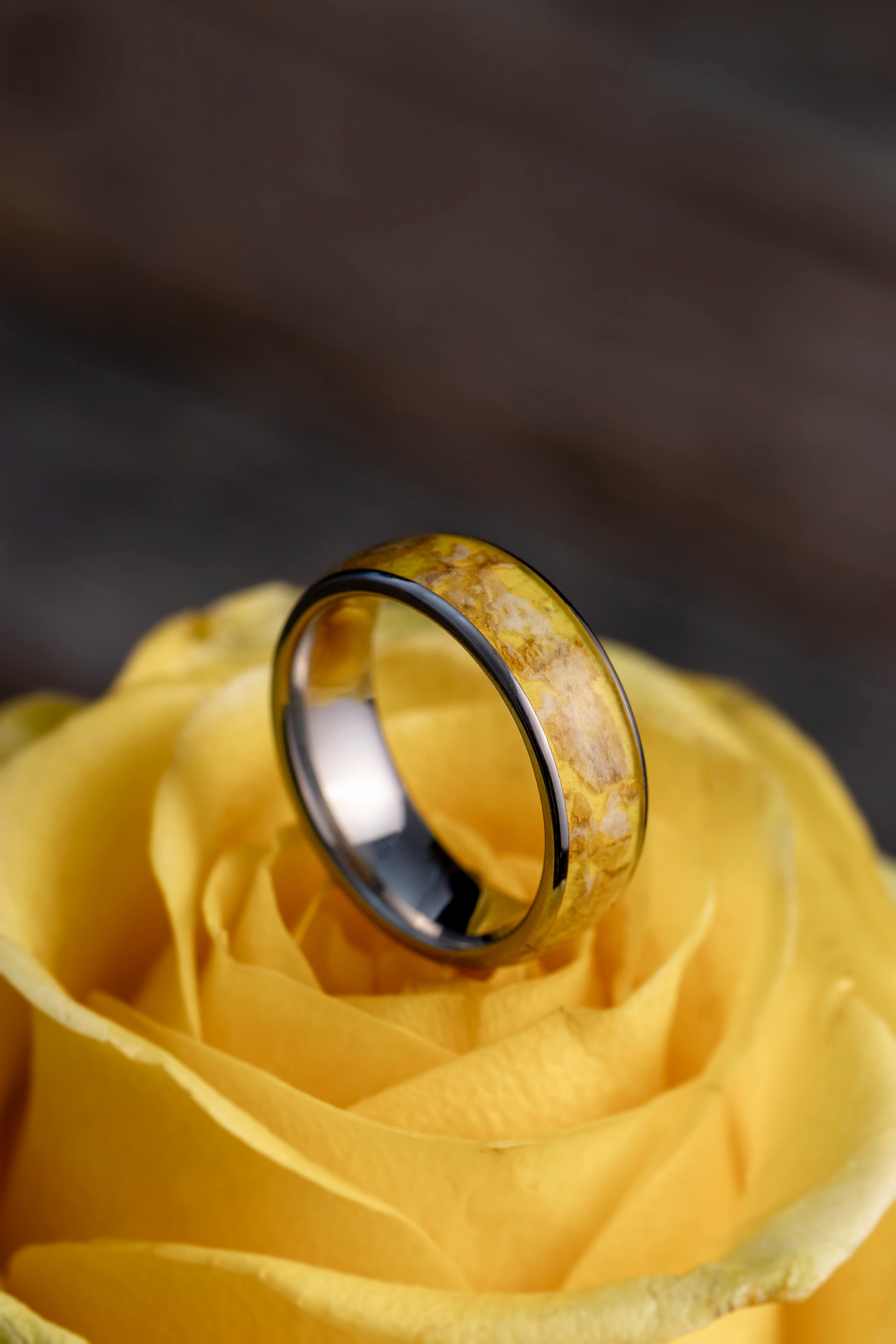 A silver ring with a yellow floral inlay rests on a yellow rose.