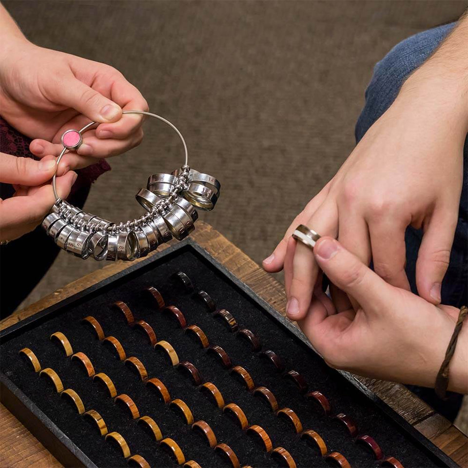 Person selecting a ring size from a set of metallic rings held by another person, with a box of various ring sizes in the foreground.