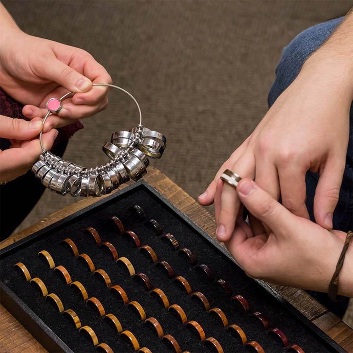 Person selecting a ring size from a set of metallic rings held by another person, with a box of various ring sizes in the foreground.
