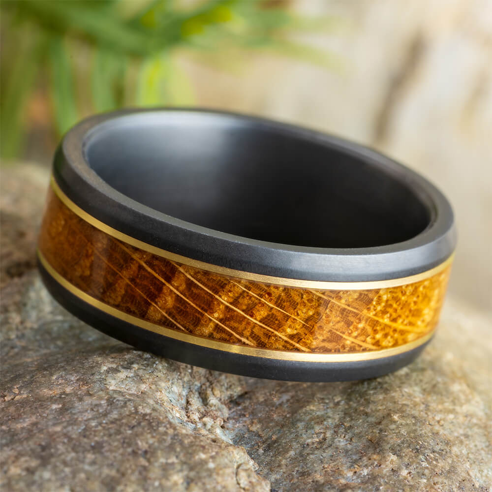 Gold and black zirconium wedding band with whiskey oak inlay, 8mm wide, featuring a beveled profile and matte finish, displayed on a stone surface.