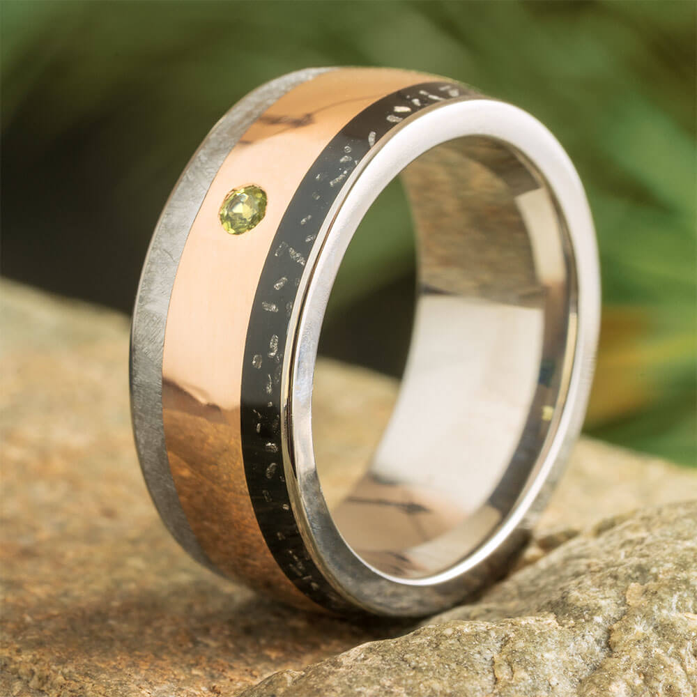 Meteorite wedding band for men featuring rose gold, black stardust inlay, and a flush-set peridot stone. Polished titanium edges add contrast.
