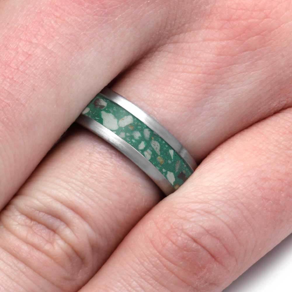A close-up of a ring worn on a finger, featuring a green inlay with white and gold flecks, bordered by a polished silver metal band.