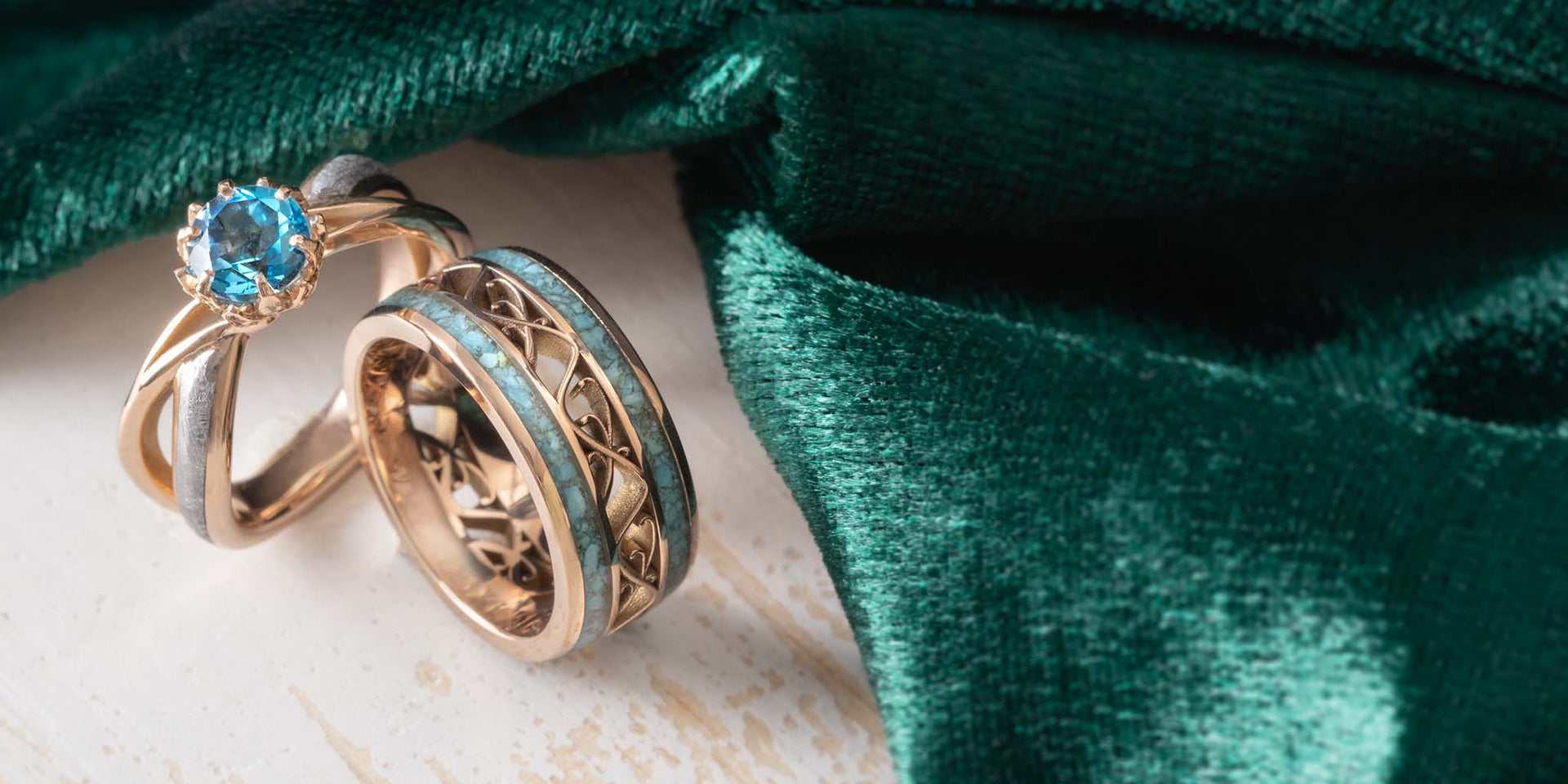 A close-up of two rings on a light surface with dark green velvet fabric behind them: a gold ring with a round blue gemstone and a gold ring with a turquoise inlay, both showing turquoise-blue inlay details.